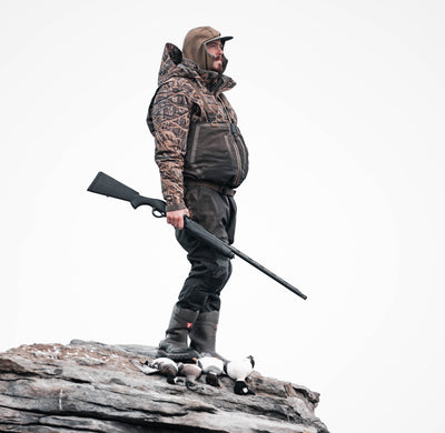 Wearing the Waterfowl LST Hardline 3-in-1 Jacket, a person in camouflage stands on rocky terrain with a rifle, three birds at their feet, set against a plain white background.