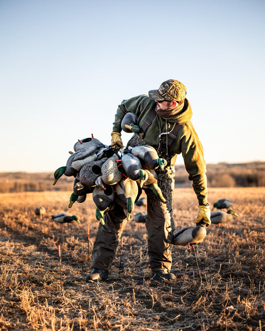 At sunrise, a person wears the Waterfowl LST CommandXT Bib with Primaloft in a field, carrying duck decoys. More decoys are scattered on dry ground as distant hills rise under a clear sky.