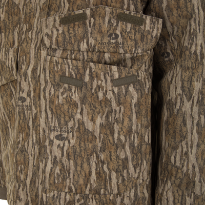 Close-up of a Spoonie Shoveler Insulated Jacket in a brown tree bark camouflage pattern with large flap pockets and Mossy Oak Bottomland logos.