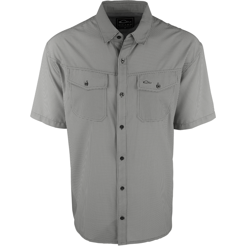 A close-up of the Traveler's Check Shirt S/S, a black and white shirt with collar and button details. Made with a lightweight, breathable poly/spandex fabric for ultimate comfort and freedom of movement. Ideal for the man on the go, whether on vacation or running errands. Moisture-wicking and wrinkle-resistant. 2 chest pockets with button flaps.