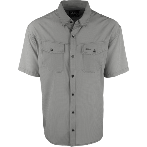 A close-up of the Traveler's Check Shirt S/S, a black and white shirt with collar and button details. Made with a lightweight, breathable poly/spandex fabric for ultimate comfort and freedom of movement. Ideal for the man on the go, whether on vacation or running errands. Moisture-wicking and wrinkle-resistant. 2 chest pockets with button flaps.